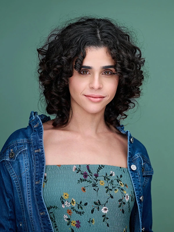 Actor headshot of a woman with curly dark hair wearing a denim jacket against a green studio background.
