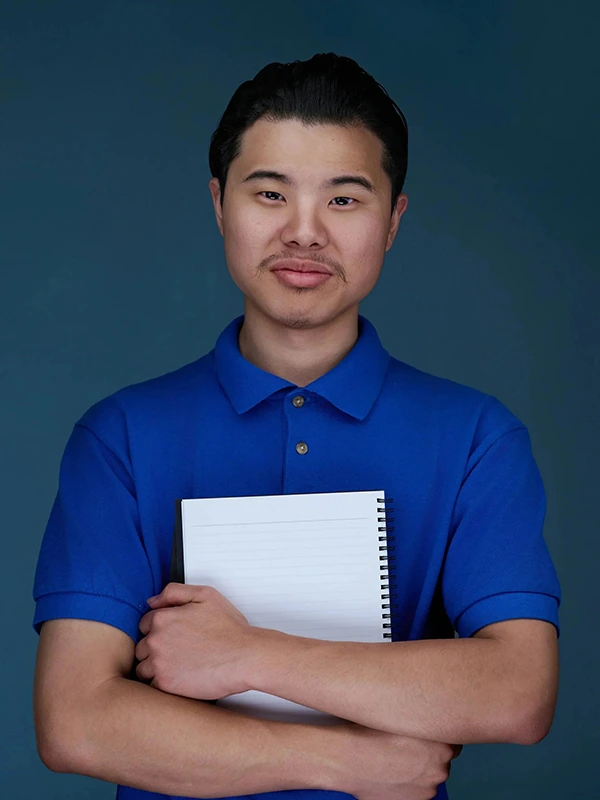 Commercial actor headshot of a young man in a blue polo holding a notebook against a blue background.