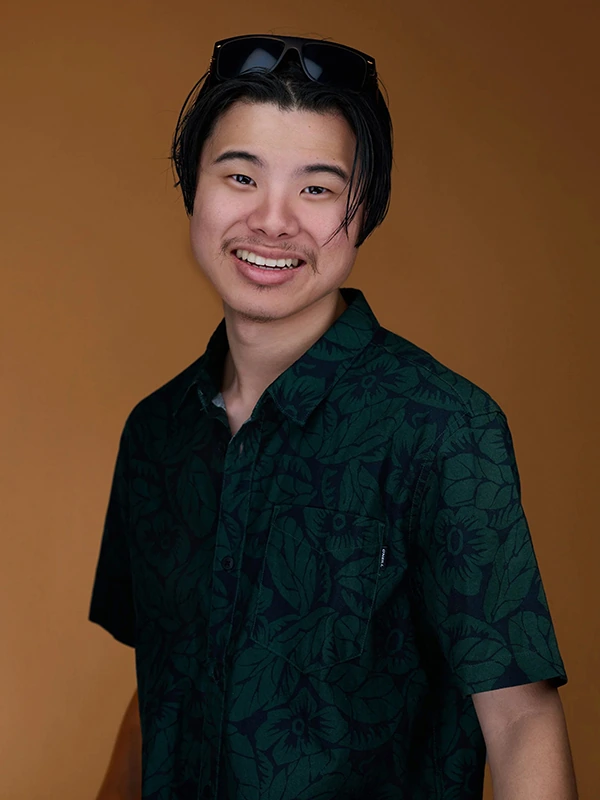 Smiling commercial actor headshot of a young man in a green patterned shirt against a warm brown background.