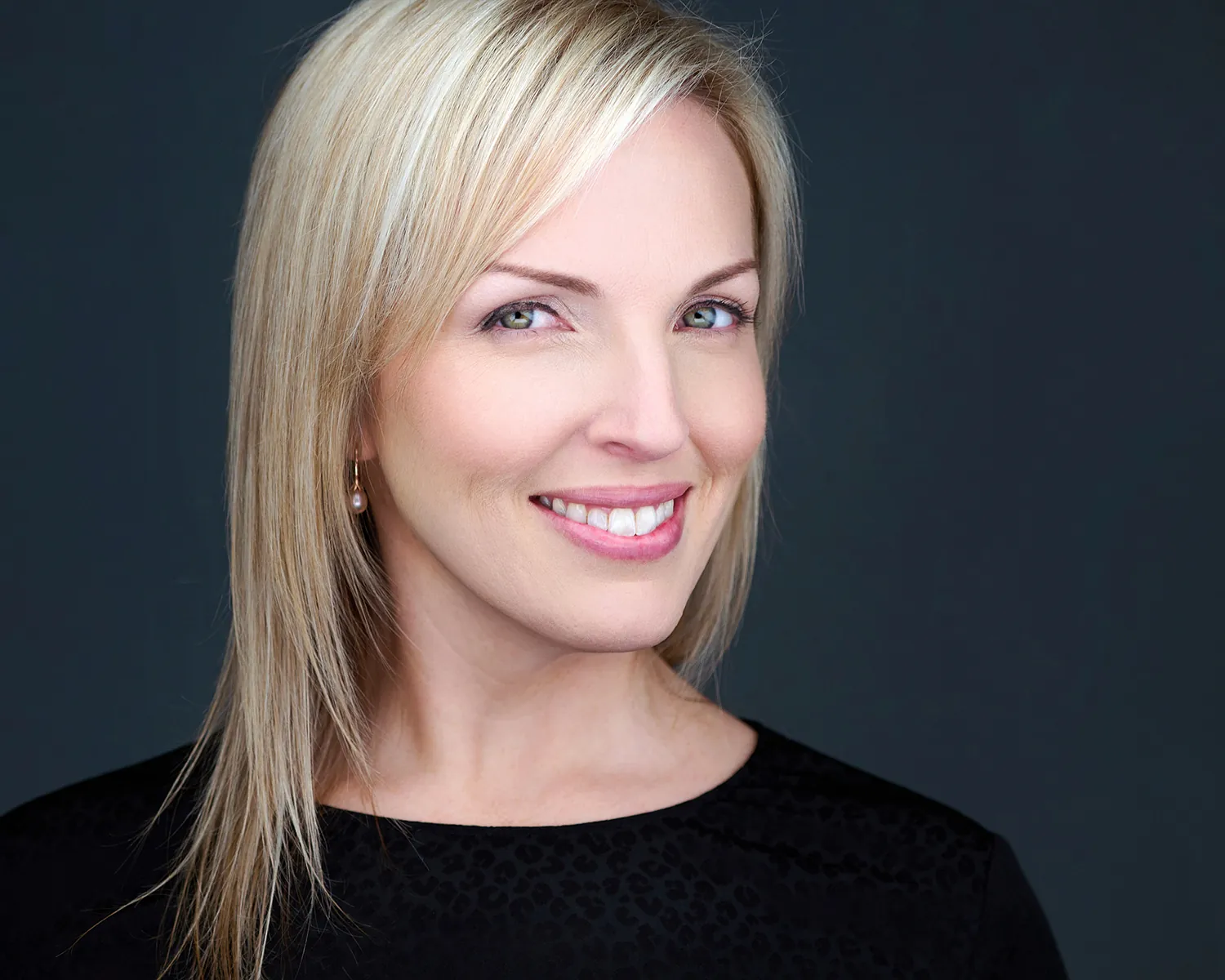 Professional headshot of a blonde woman in a black top against a dark studio background.