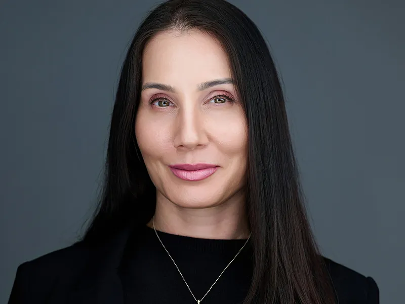 Professional headshot of a dark-haired woman in a black top against a gray background.
