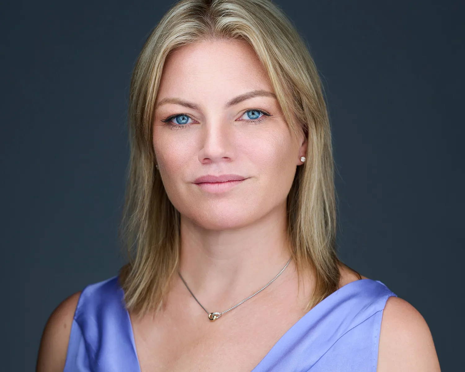 Professional headshot of a blonde woman in a lavender top against a dark studio background.