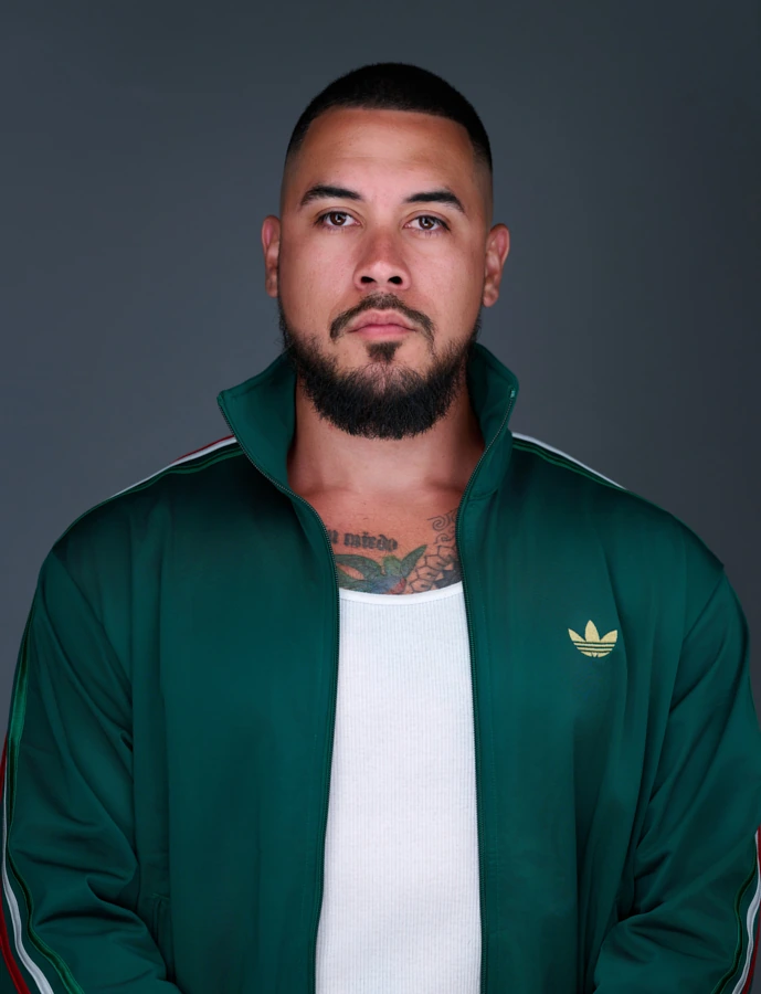 Theatrical actor headshot of a bearded man in a green track jacket against a dark gray background.