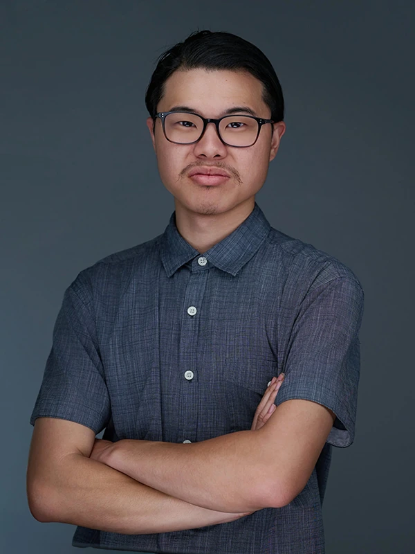 Theatrical actor headshot of a man wearing glasses and a gray shirt against a dark background.
